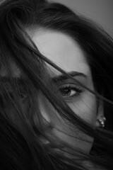 Close-up beautiful woman with seductive look studio portrait. Model covering part of her face with hairs by blowing wind. Mysterious and sensual look. Eye is in camera focus. Black and white image