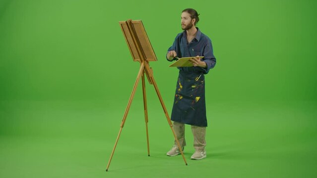 Full Height View.Young Artist in Denim Shirt and Apron Working on Creation of New Painting.Bearded Man Draws with Brush on Canvas on Easel on Green Screen,Chroma Key.The Creativity and Art Concept