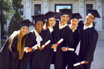 Laughing excited group of multicultural people in graduation gowns caps outdoors in campus. They stand leaning to the right. Group portrait. Graduation from high school, college, university concept.