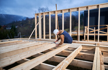 Man worker building wooden frame house on pile foundation. Carpenter hammering nail into wooden plank, using hammer. Carpentry concept.