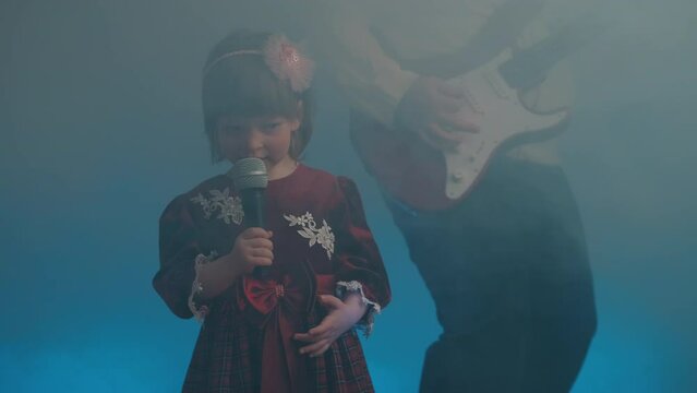 Little girl on stage in vintage dress, she sings into microphone and dances, her father plays an electric guitar. Color music is shining and smoke is billowing. Family time, creativity and hobbies