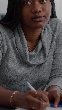 Vertical video: Portrait of student with black skin writing creative communication project for high school homework sitting at desk in living room. Teenager woman browsing graphic information on