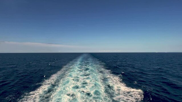 Stern view of wide water wake left from cruise ship on sea water surface with horizon in background and shadow of people leaning over parapet of deck
