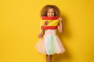 Cute little girl holding Spanish flag standing isolated over yellow background.