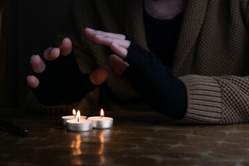 Man in sweater and fingerless gloves warming up his hands with tealight candles during energetic crisis in Europe. - obrazy, fototapety, plakaty