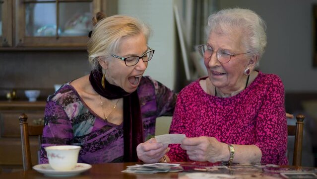 Slow motion of elderly woman and mature daughter having fun and laughing looking at old photo memories at the dining room table.