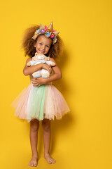 Cute child girl in elegant tulle dress and unicorn headband celebrates birthday party holding teddy bear over yellow background.