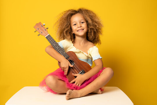 Cute little girl in summer clothing playing ukulele over yellow background. Happy vacation concept.