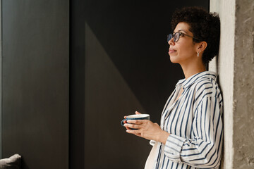 Happy businesswoman drinking coffee while standing by wall at home