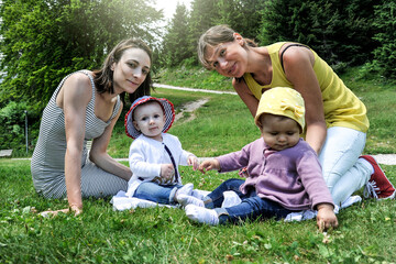 Naklejka na ściany i meble Female lesbian couple with two adopted baby girl during a pic-nic at the park - Two mothers relaxing with the baby girls on lawn - Female babysitters - LGBT family and diversity concept