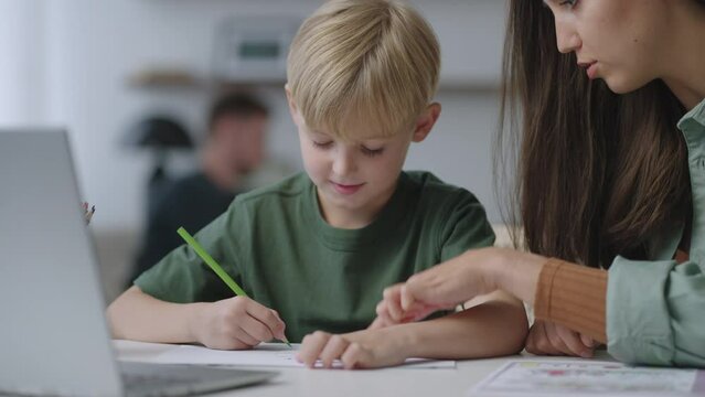 Portrait :A family with two children, dad plays with his daughter, and mom helps her son do his homework, explains the topic and talks to the boy sitting at the table in the living room