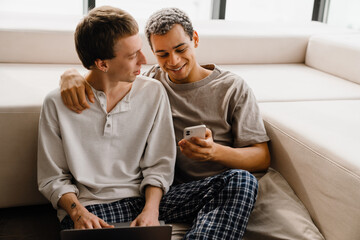 Happy gay couple using cellphone and laptop while resting at home