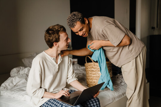 Happy Young Gay Couple Using Laptop And Folding Clothes In Bedroom