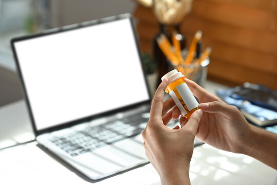 Close up image of a woman's hand holding a pill bottle to read the drug label with the laptop with empty screen as the background.