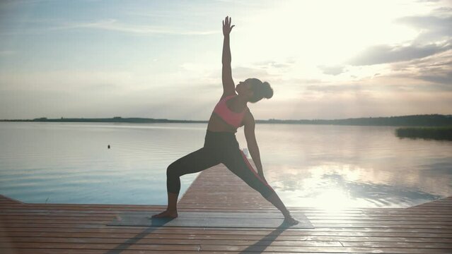 Side view of beautiful African American young sporty female in sportswear practice yoga on lake on dock on sunrise or sunset. Woman athlete working out on nature standing in yoga warrior posture