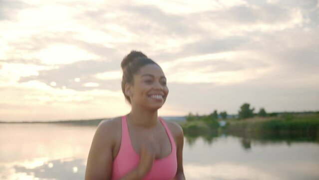 Close Up Of Joyful Smiling African American Woman Athlete Working Out Outdoors Running Near Lake On Sunset. Beautiful Young Female In Sport Top Runs On Nature With Beautiful View. Sporty Lifestyle