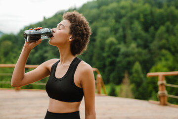 Curly woman drinking water during yoga practice in mountain