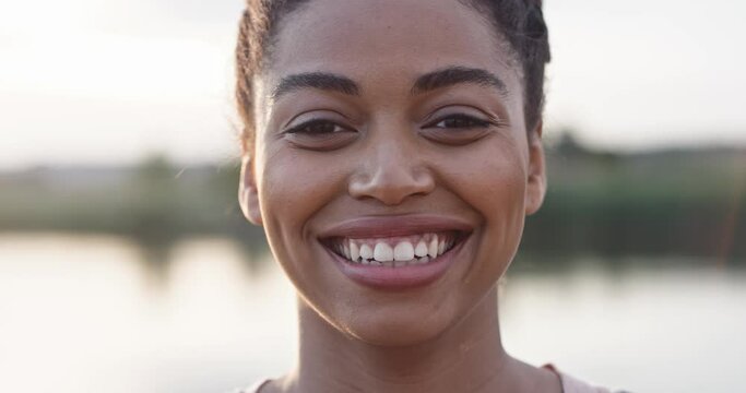Close up of face of beautiful young African American woman looking at camera and smiling outdoors in nature. Lake on blurred background. Happy female talking and laughing. Positive emotions