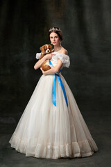 Beloved pet. Portrait of young charming woman in white vintage dress as medieval princess holding small King Charles spaniel dog on dark background. © master1305