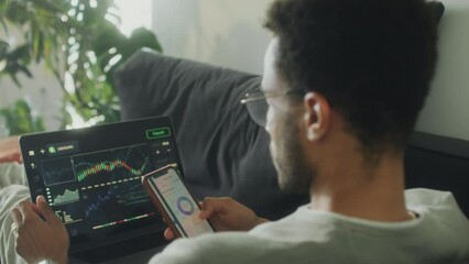 Over the shoulder shot of young man sitting on sofa at home, analyzing stock market graph on laptop and using finance app on phone while trading online - Powered by Adobe