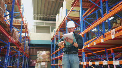 Caucasian logistics employee is holding paperwork to examine the stock of cargo cardboard boxes. Young adult logistics engineer with responsibility to manage the stock of goods in a large warehouse.