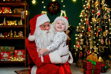 Fototapeta na wymiar Laughing little boy in Santa's hat hugging with Santa Claus near Christmas tree. New Year concept 