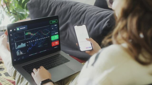 Over the shoulder shot of girl sitting on couch, using finance app on smartphone and analyzing stock market chart on laptop while trading online from home