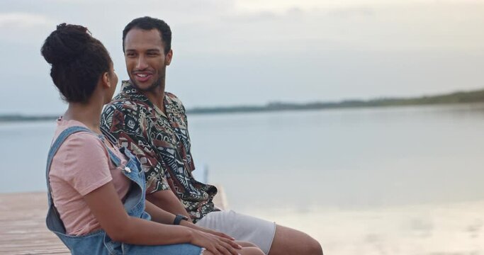 Close up of happy smiling mixed-race couple sitting on dock looking at each other and chatting. Boyfriend and girlfriend spending time together near water. Weekend rest. Love relations, romantic