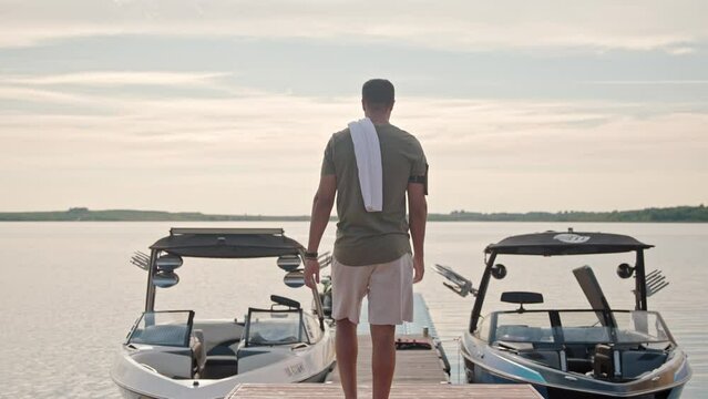 Back view of man in shirt and shorts with towel on shoulder walking on the dock between boats on lake. Rear view. One young male going on pier outdoors near two yachts on water. Sport and recreation