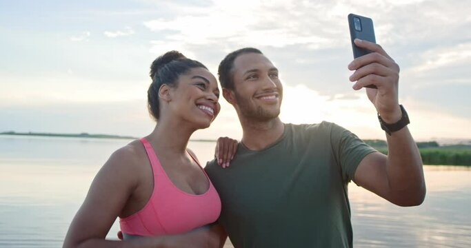 Joyful positive young couple in love man and woman athletes standing on lake posing to smartphone camera smiling and hugging. African American family couple taking selfie photo on mobile phone