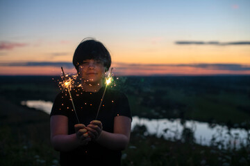 A cute boy holds sparklers in his hands, fireworks in the evening in the summer at sunset. Festive mood.