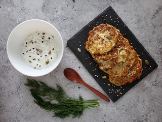 Zucchini fritters, sour cream sauce in white bowl, dill sprig, wooden spoon on light background. 