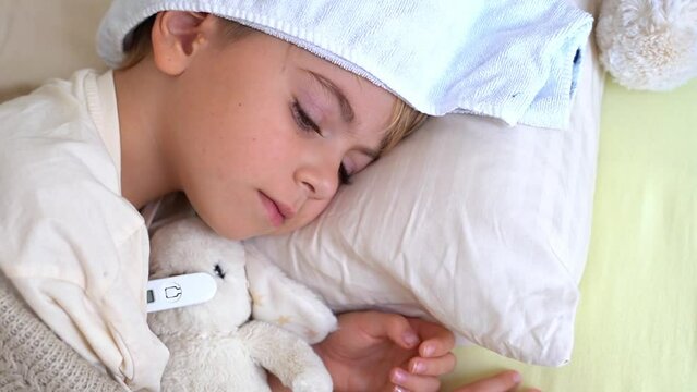 Sick fever child lying in bed with cold compress on his forehead, measure body temperature with electronic thermometer. Children cold and flu, coronavirus illness concept. Top view.