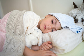 Sick fever child lying in bed with cold compress on his forehead, measure body temperature with electronic thermometer. Children cold and flu, coronavirus illness concept.