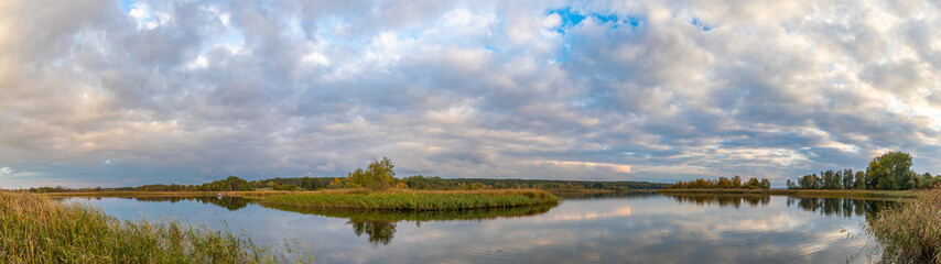 Obraz premium Landschaft mit Bäumen, Schilf, wolkigem Himmel und der Spiegelung der Wolken im See