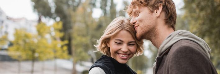 happy redhead man kissing head of blonde girlfriend smiling in park, banner.