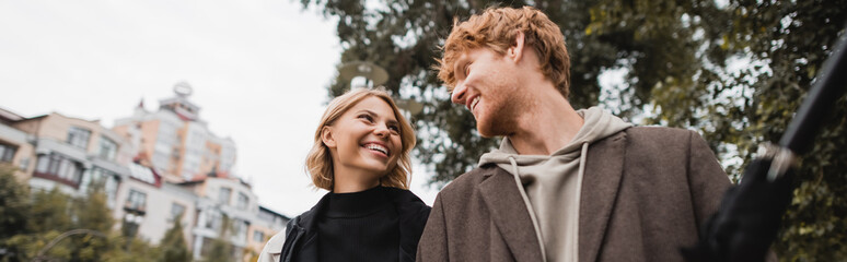 happy redhead man and joyful blonde woman looking at each other while walking in park, banner.