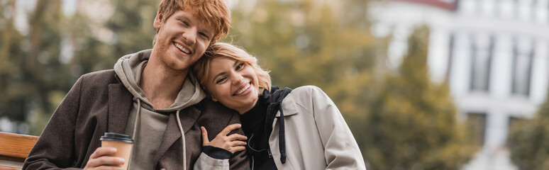 Obraz premium happy young woman hugging boyfriend holding paper cup with coffee to go in park, banner.