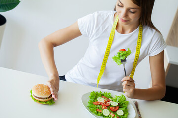 Beautiful young woman decides eating hamburger or fresh salad in kitchen.