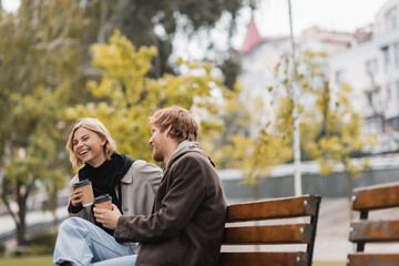 joyful young couple laughing and holding paper cups with coffee to go while sitting on bench in...