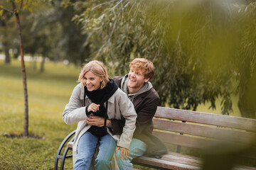 redhead man smiling while tickling positive girlfriend while sitting on bench.