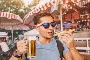 Happy man drinking beer and eating traditional german bratwurst - hotdog at funfair and street food...