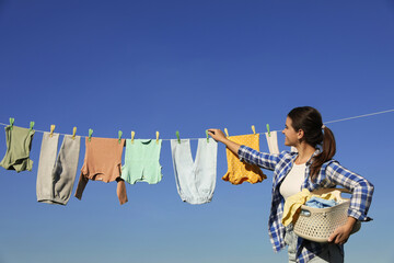 Smiling woman hanging baby clothes with clothespins on washing line for drying against blue sky...