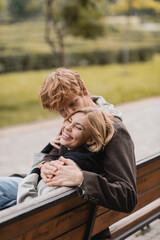 redhead man hugging and kissing positive woman while sitting on wooden bench in park.
