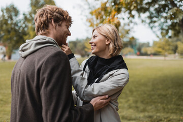 pleased couple in coats looking at each other while hugging in autumnal park during date.