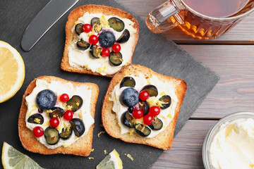 Tasty sandwiches with cream cheese, blueberries, red currants and lemon zest near cup of tea on wooden table, flat lay