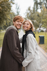 Fototapeta premium positive blonde woman and cheerful redhead man holding hands while strolling in park.