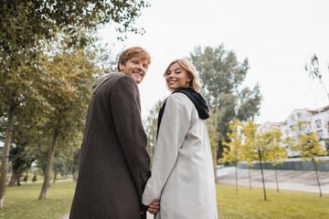 low angle view of joyful blonde woman and cheerful redhead man holding hands while strolling in park.