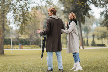 Fototapeta premium full length of happy young man in coat holding paper cup and standing with girlfriend in autumnal park.