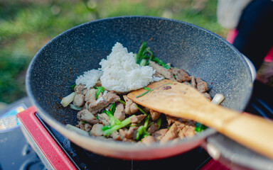Chef is stir frying rice, pork, spring onions in black pan on gas picnic stove, camping menu.